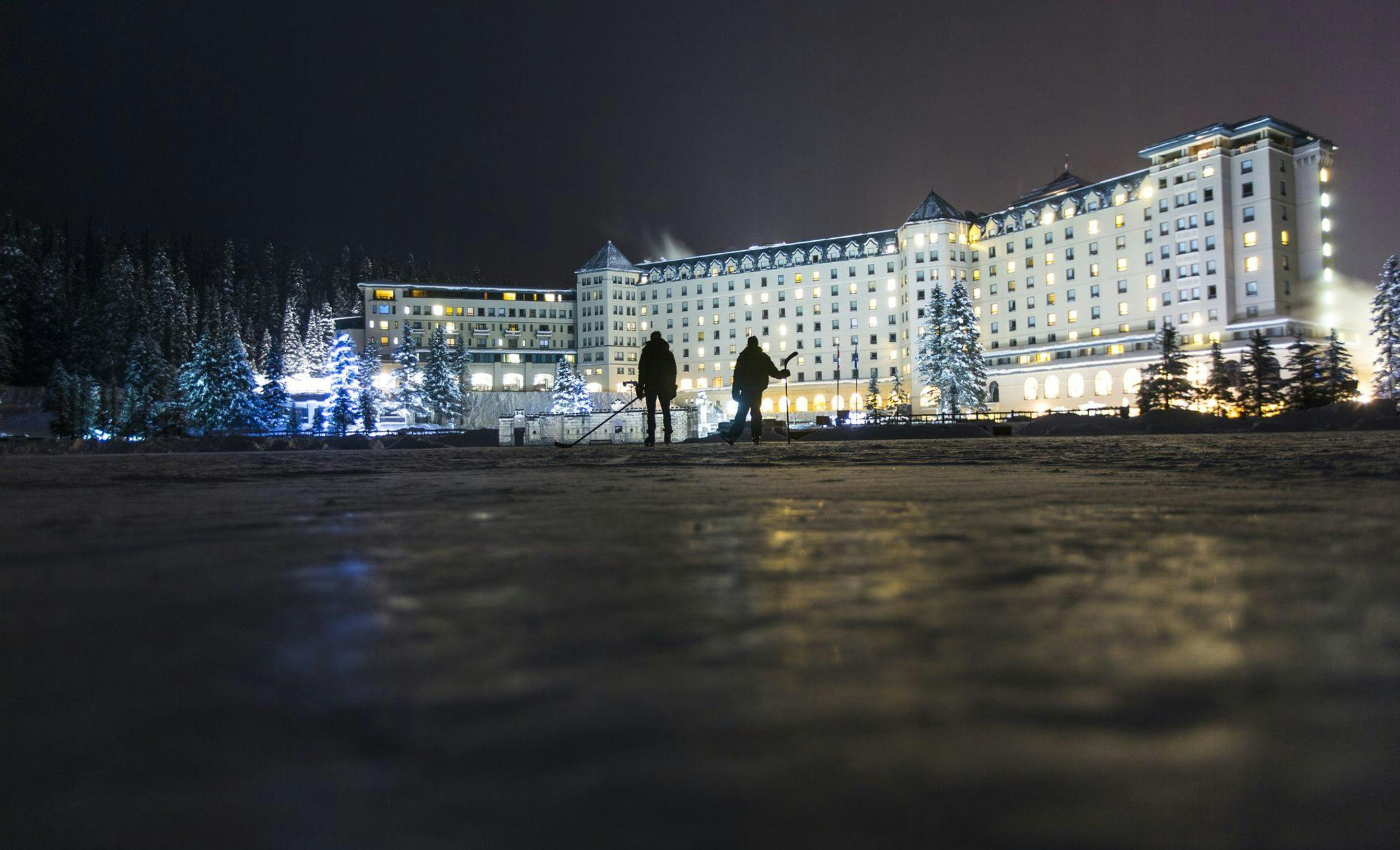 Two figures are illuminated skating on a frozen lake with a large and well lit hotel in the background. Christmas lights can be seen along the lakeshore