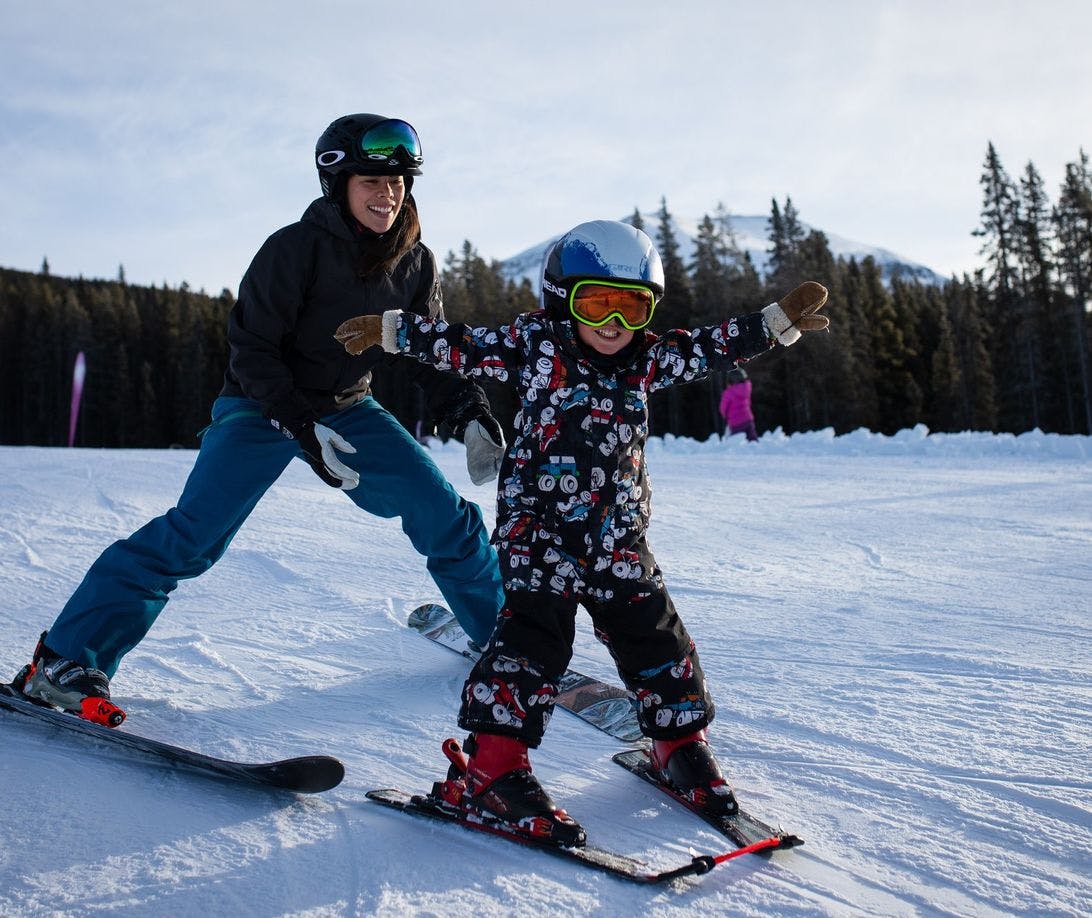 A mom skis closely behind her child as they descend a small snowy hill at a ski resort