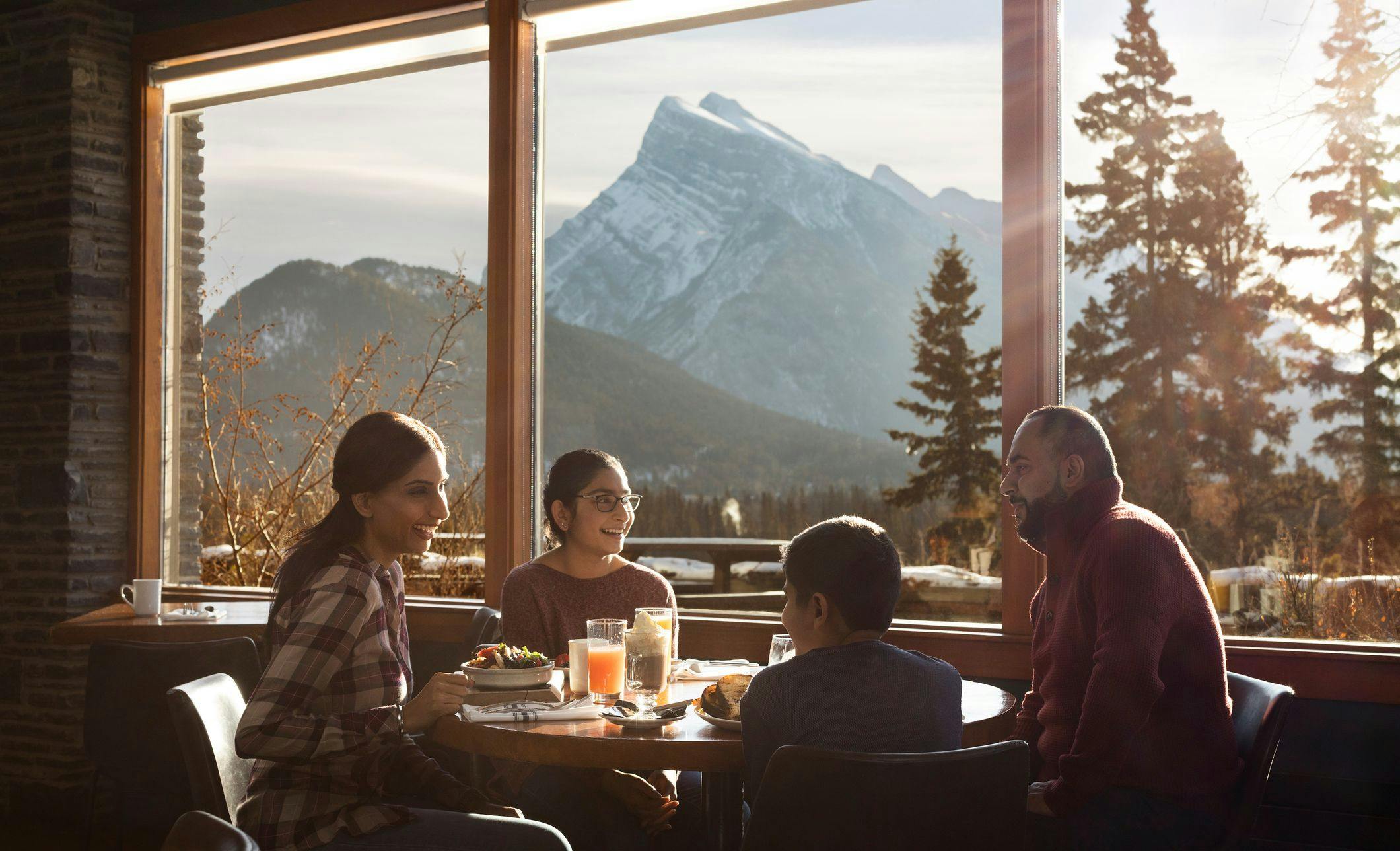 A family of four sit around a table laughing and enjoying brunch with views of a large mountain outside the window on a sunny day