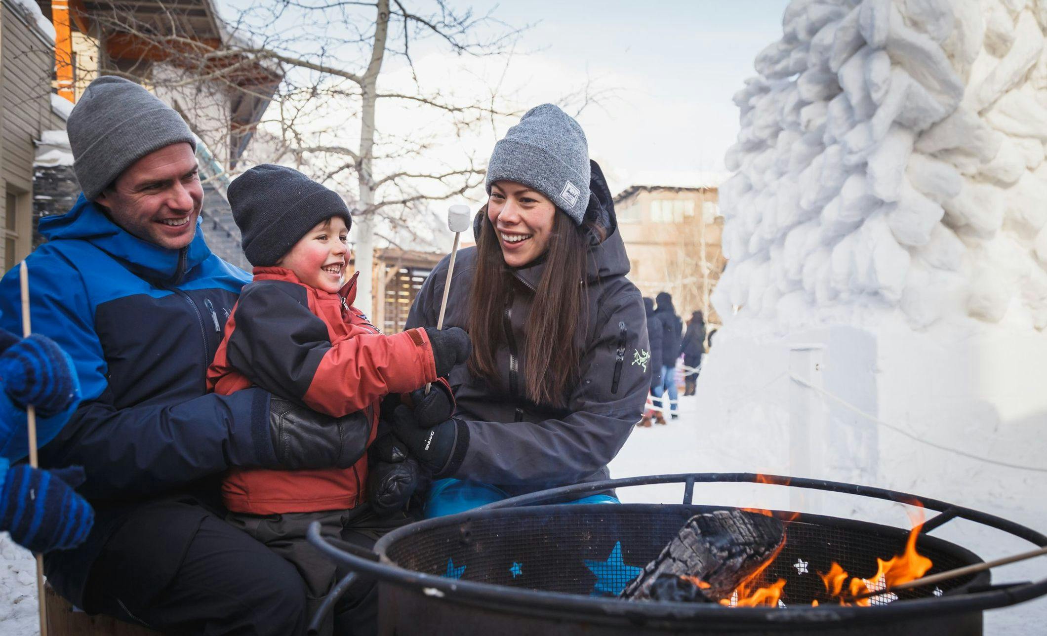A family wearing lots of cozy winter layers roasting a marshmallow at an outdoor fire pit on a snowy street