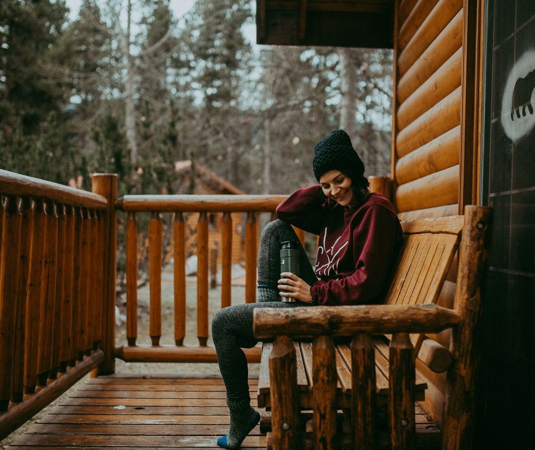 Woman sitting outside a cozy, wooden cabin wearing sweats, a toque, and drinking coffee