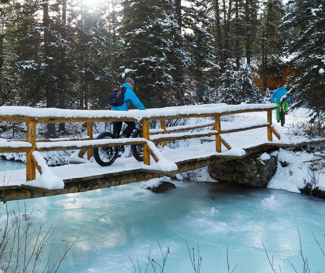 Two cyclists riding fat bikes across a snow covered bridge and over a frozen river
