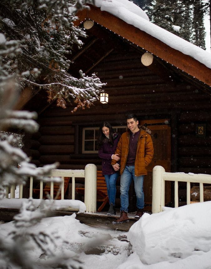 A couple in front of a cozy winter cabin surrounded by snowy trees in Banff National Park.