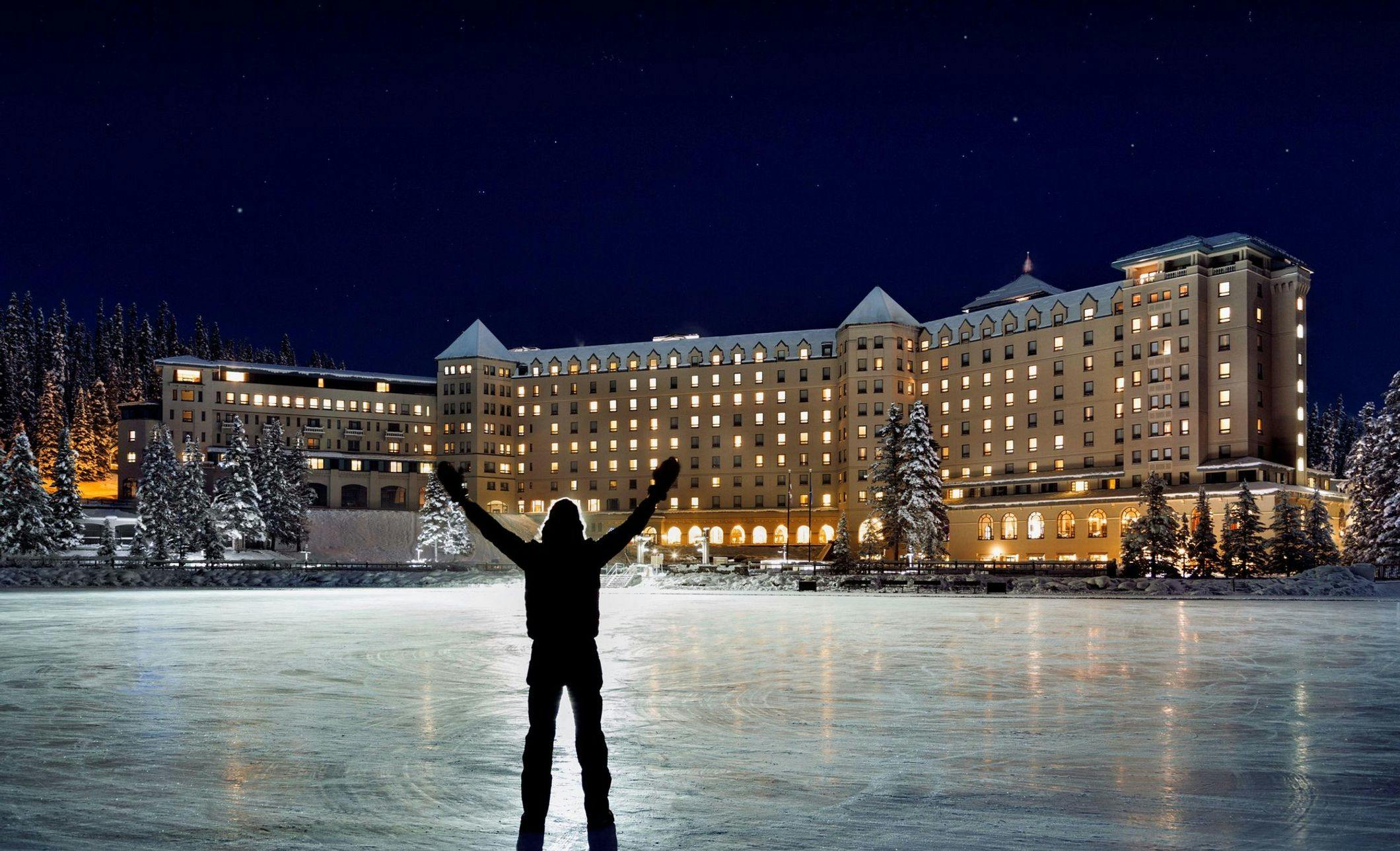 A skater stands on a lit outdoor natural skating rink at night with a large, well lit hotel next to the skating rink