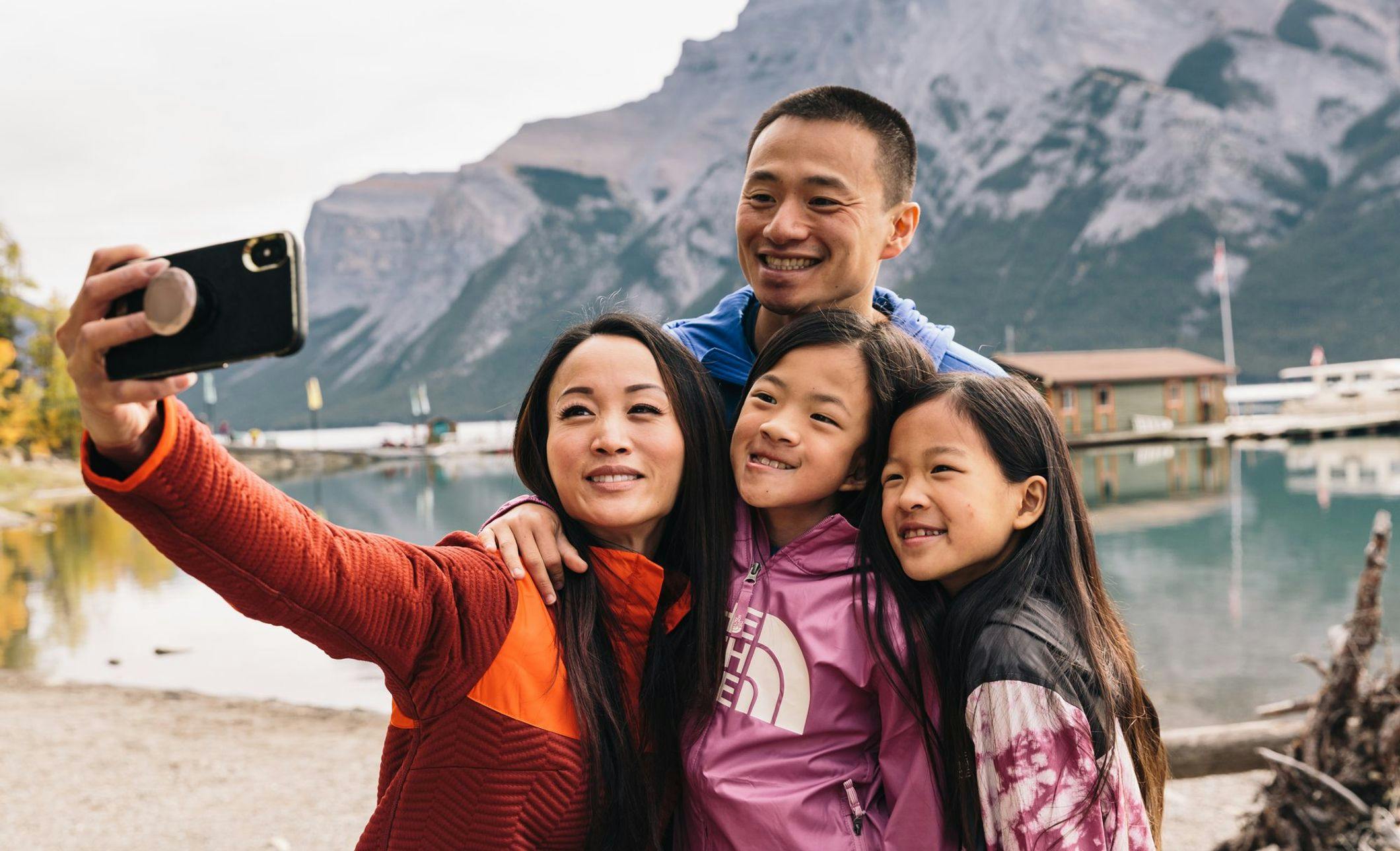 A family of four stops to take a selfie in front of a turquoise blue lake while dressed for time in the outdoors