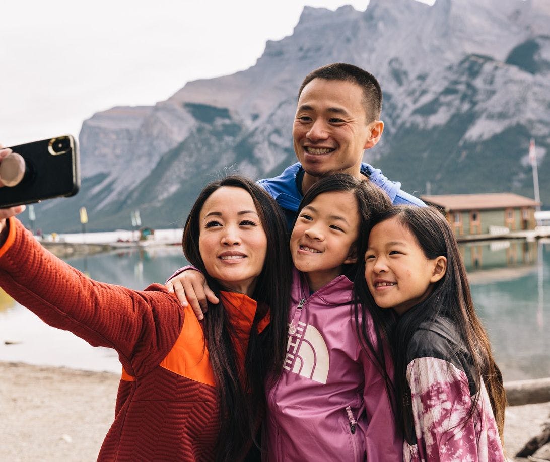 A family of four stops to take a selfie in front of a turquoise blue lake while dressed for time in the outdoors