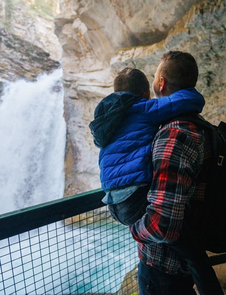 A father wearing a red flannel shirt holds his son in a bright blue jacket as they look at a rushing waterfall from behind the safety of a guardrail
