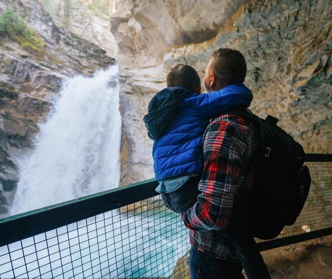 A father holds his son next to a railing overlooking at the lower waterfall at Johnston Canyon in Banff National Park. They are wearing layers in the crisp spring weather