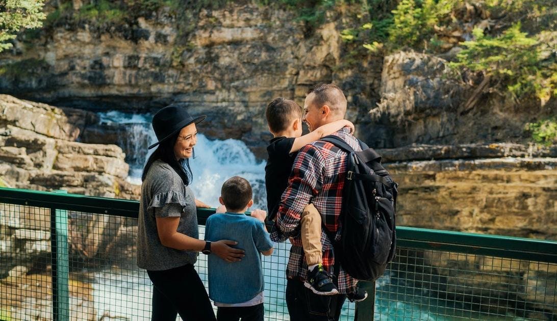 A family of four stands at a railing overlooking a rushing waterfall as they laugh and embrace each other
