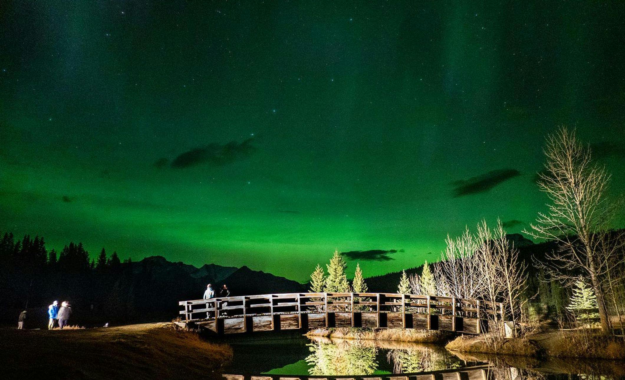 A bridge over a river at night with green aurora lighting up the sky and people out admiring it