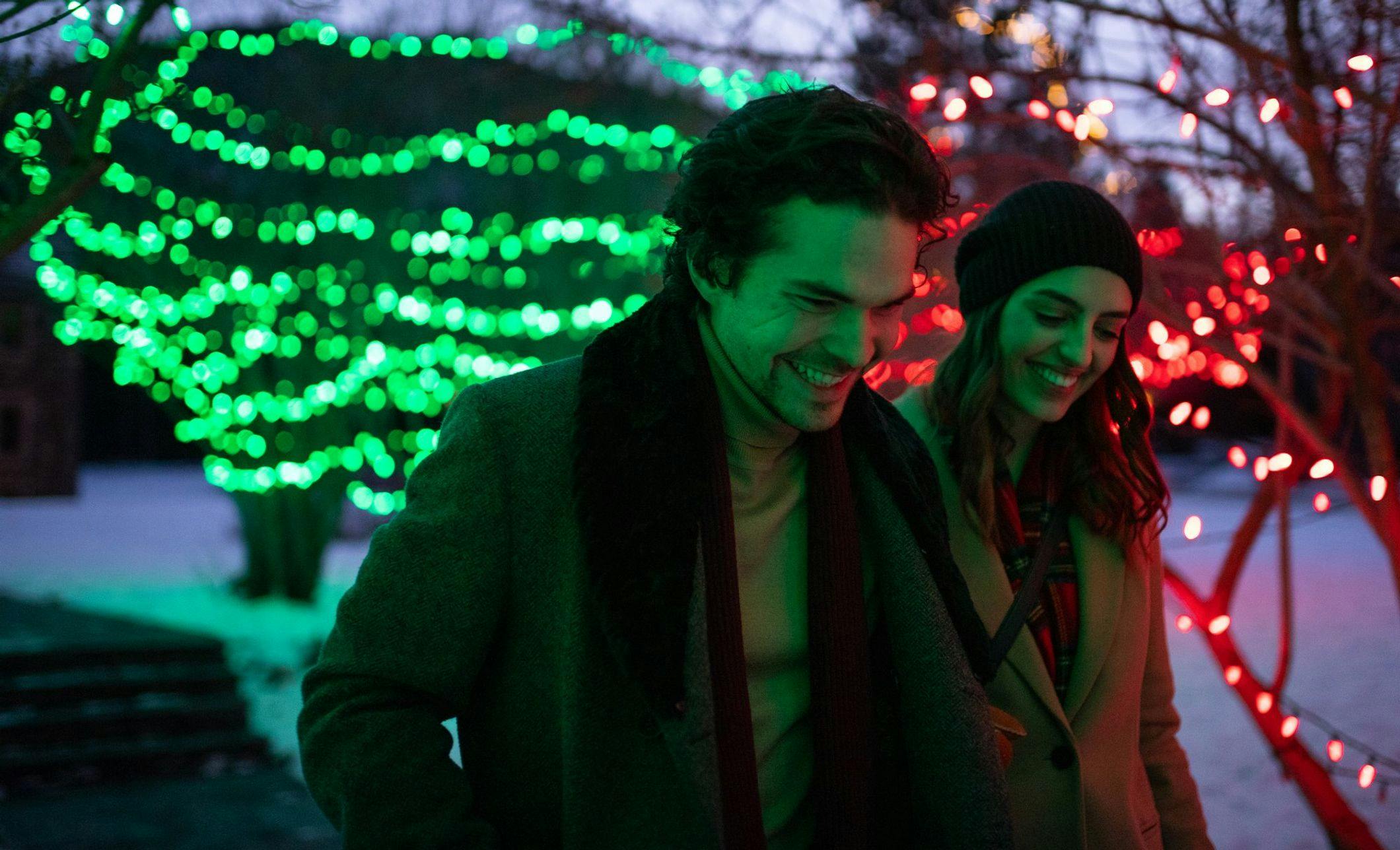 A couple enjoys an evening walk on a snowy trail surrounded by trees adorned with Christmas lights