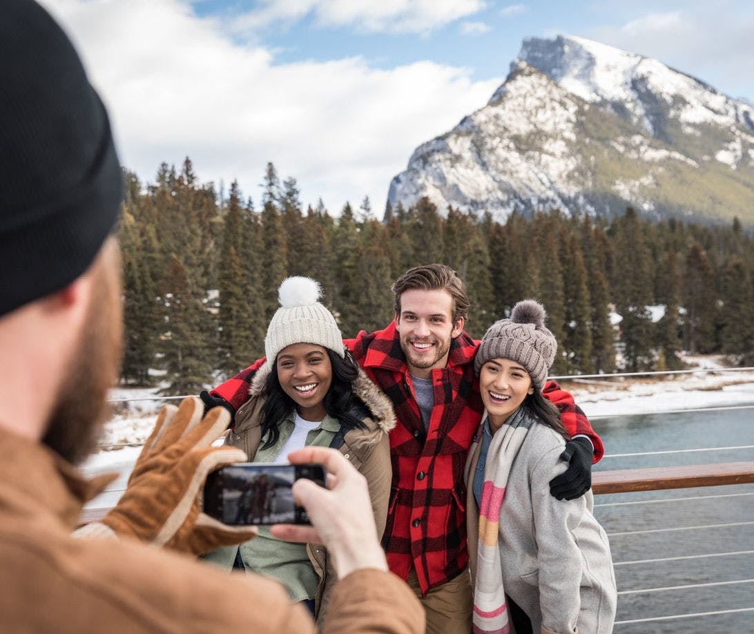 Three friends pose for a photo wearing light winter layers and a snowy Mount Rundle behind them