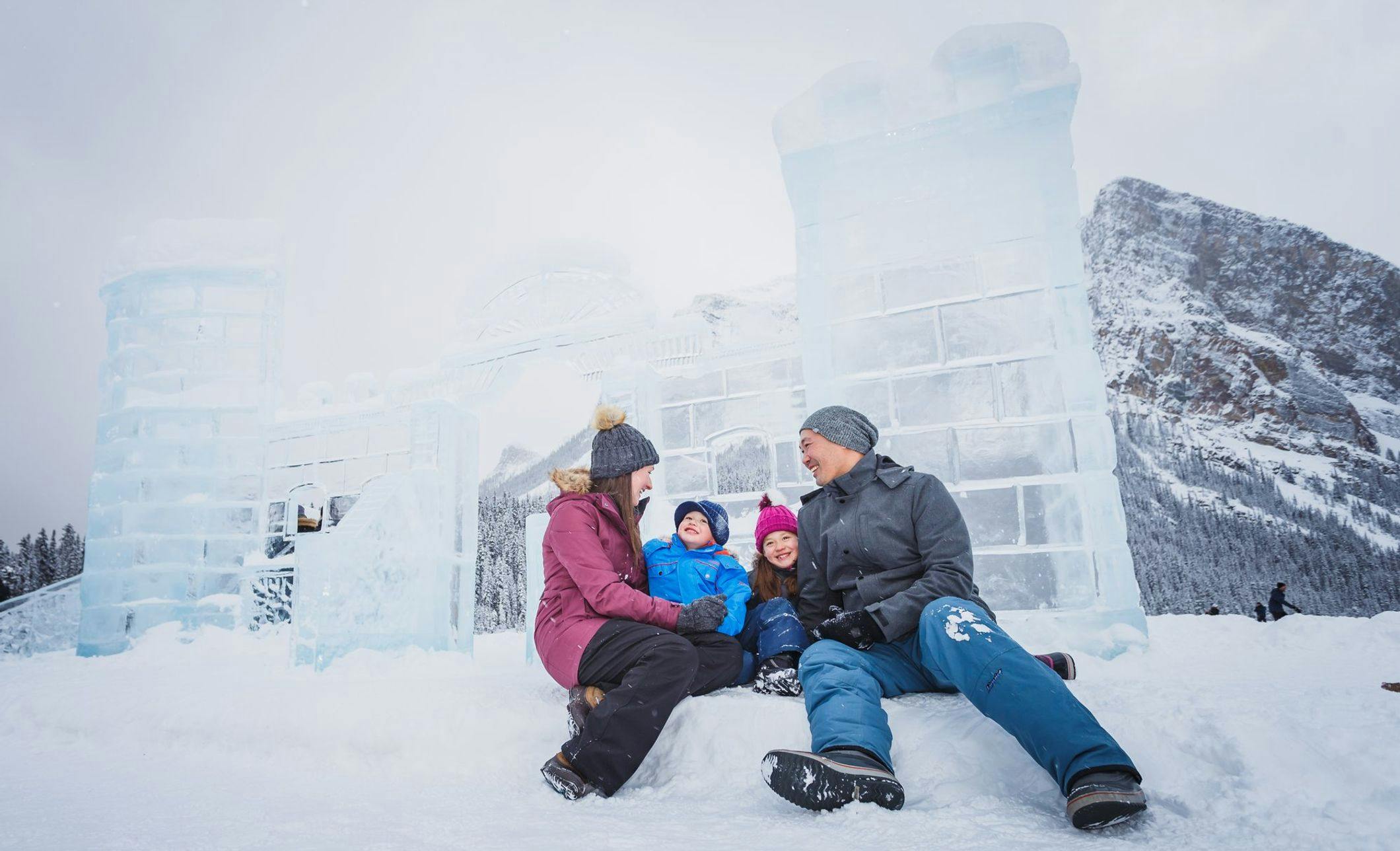 A family of four wearing all their snow gear sitting in front of a giant ice castle