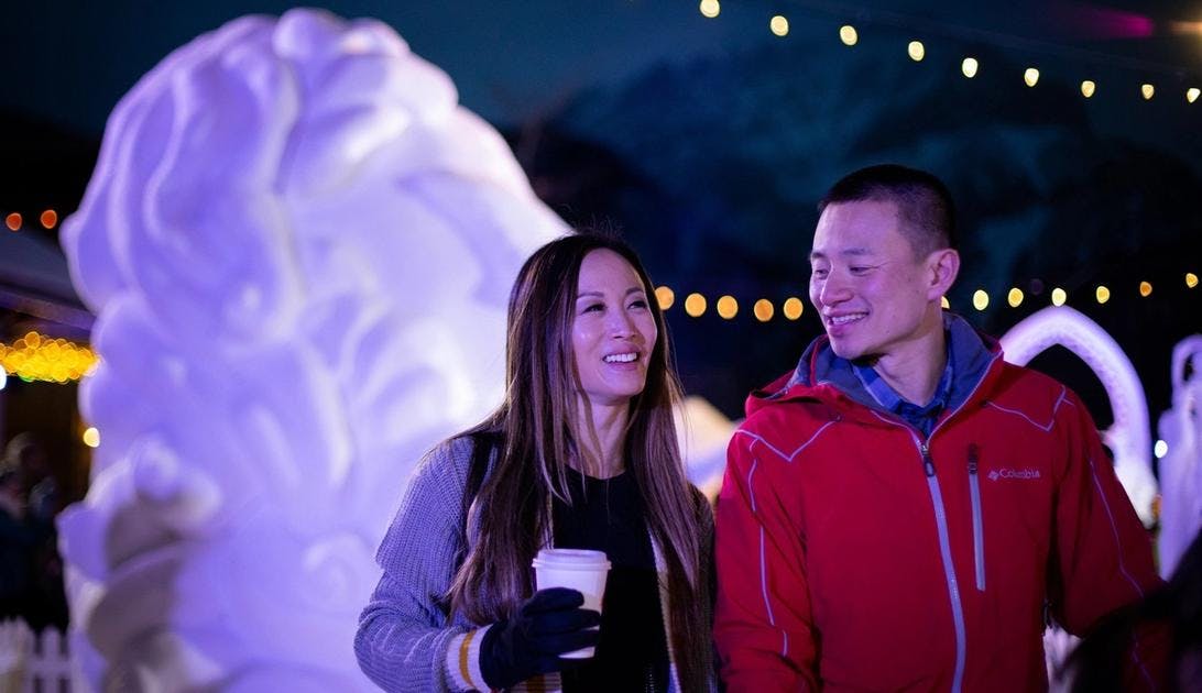 A couple walking in front of a giant snow sculpture with string lights above them in the evening