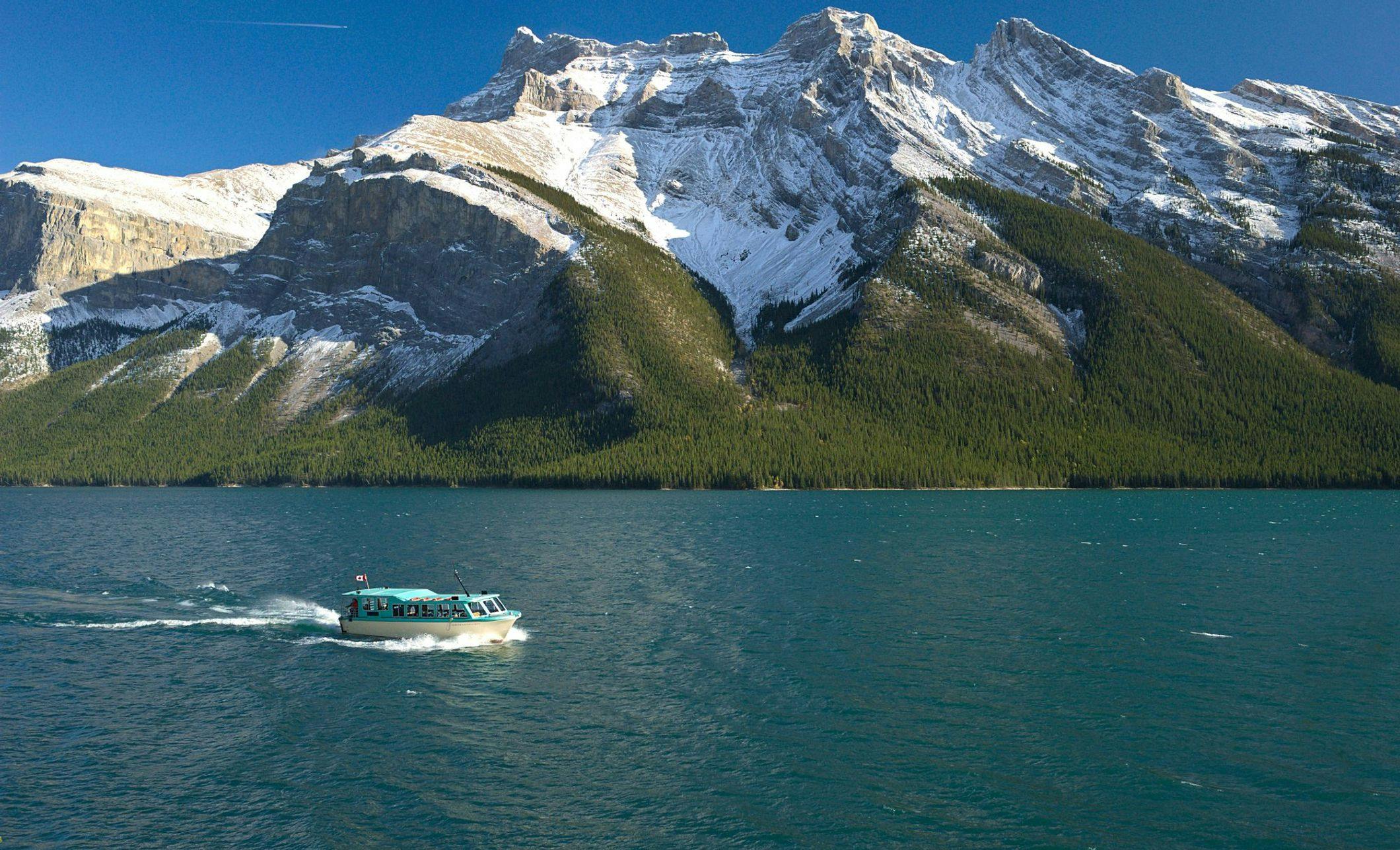 A covered sightseeing passenger boat sails along vibrant blue water with a snow-capped mountain in the background