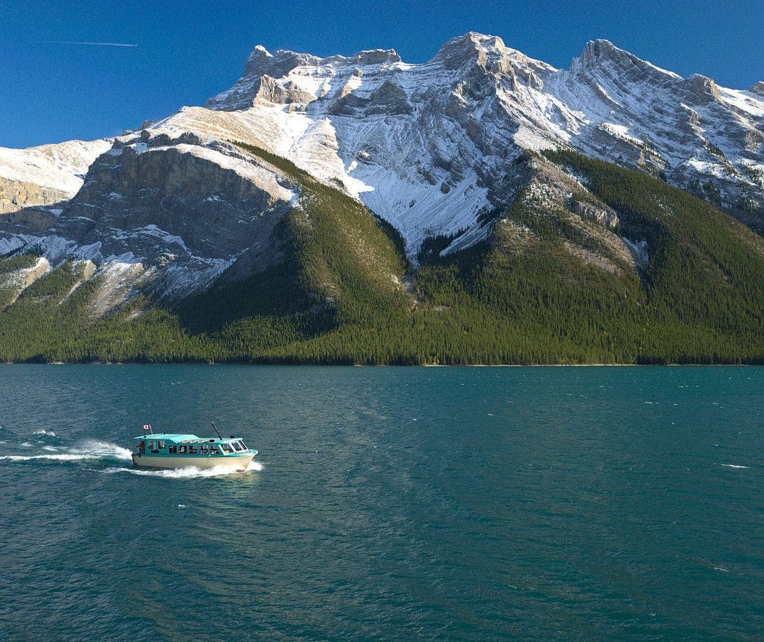 A small cruise boat travels along a vast blue lake with tall mountain peaks surrounding