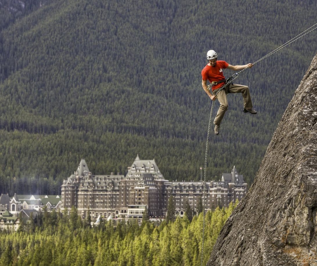 A climber hangs mid-air while attached to the rockface of a mountain with the Fairmont Banff Springs Hotel in the background