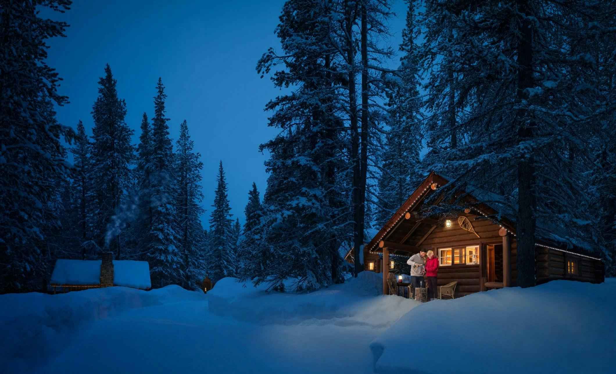 Cabin surrounded by snow in the evening with a couple on the porch sharing a bottle of wine