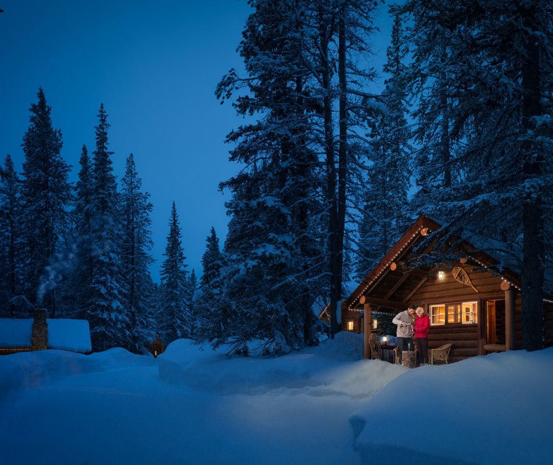 Cabin surrounded by snow in the evening with a couple on the porch sharing a bottle of wine