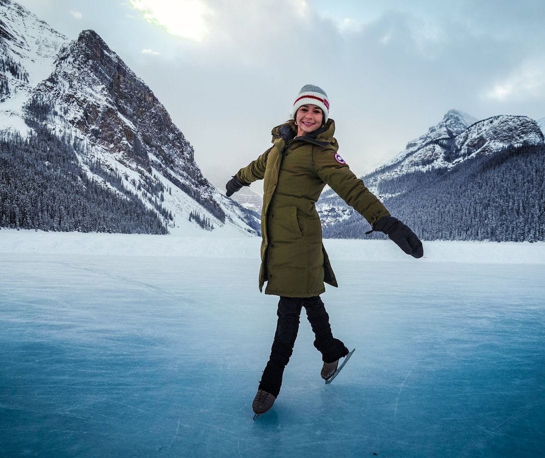 A woman poses while skating on a large frozen lake framed by mountains and a glacier behind her