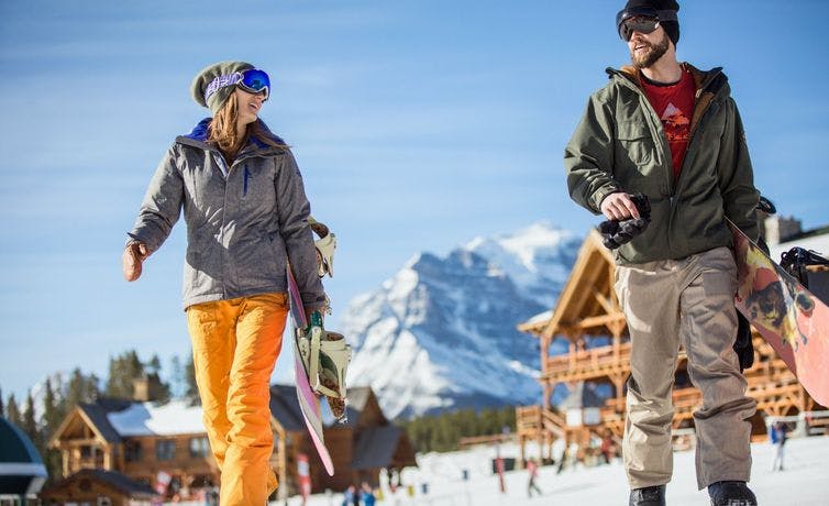 A man and woman walking across a snowy ski resort with the lodge in the background. Both are wearing their goggles as it is a bright sunny day