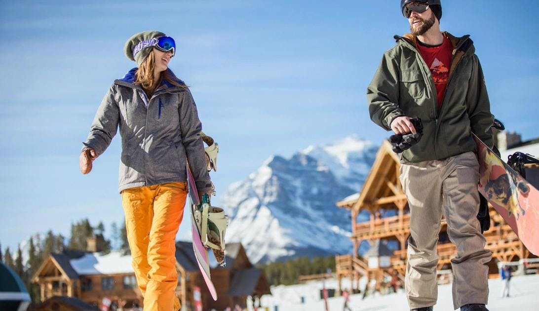 A man and woman walking across a snowy ski resort with the lodge in the background. Both are wearing their goggles as it is a bright sunny day