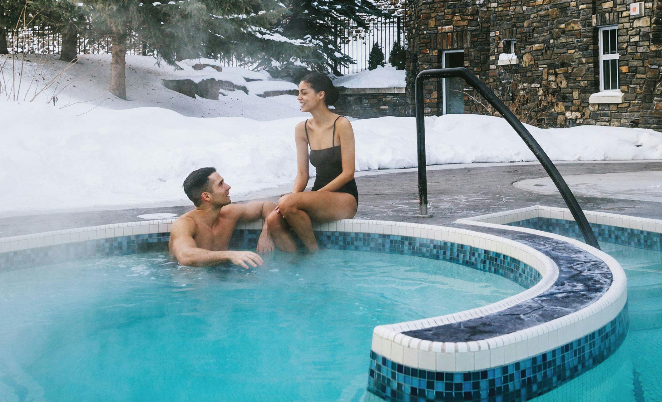 A couple soaking in an outdoor hot tub with a snowy background behind them and steam rising in front of them