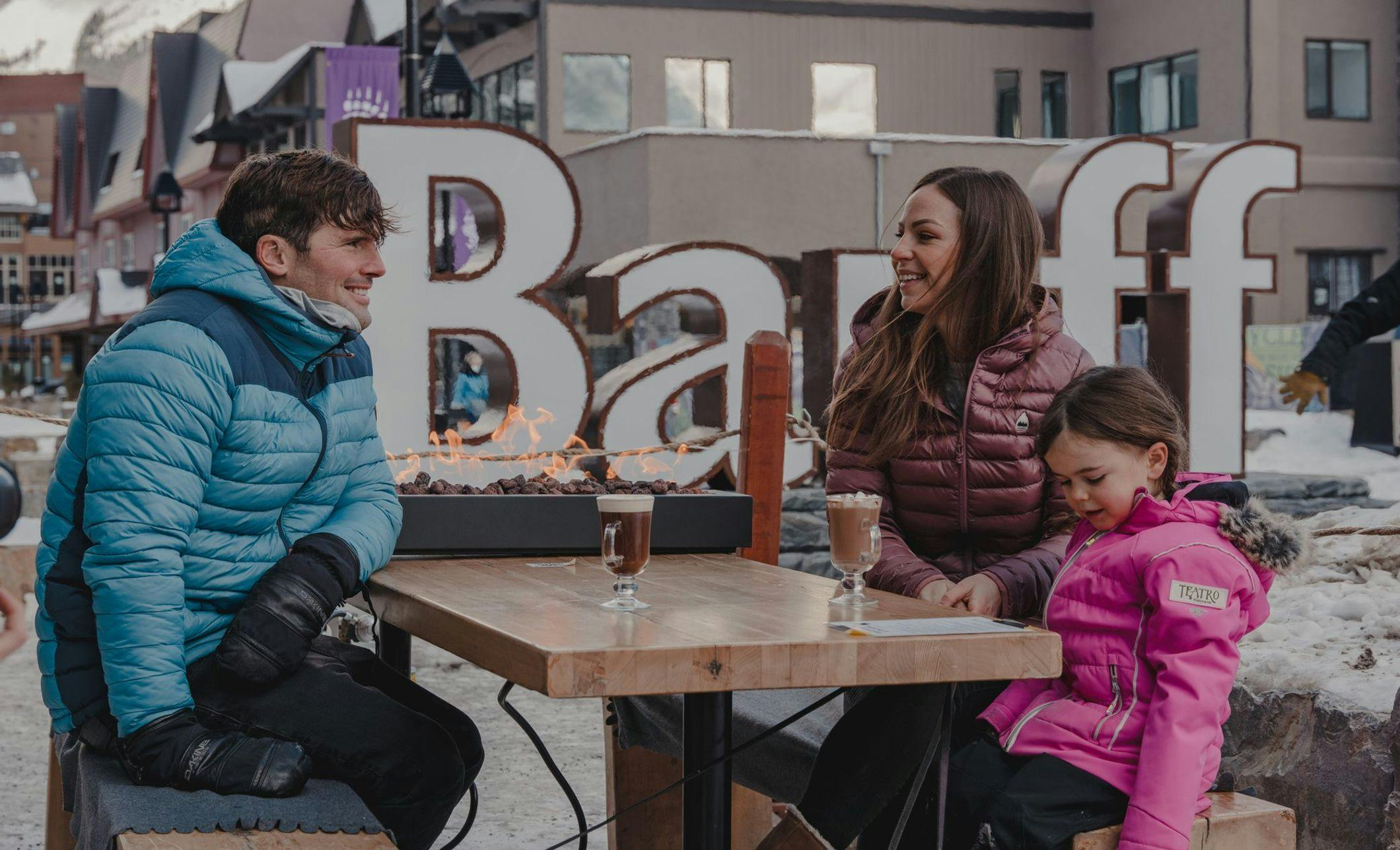 A family of three sit at an outdoor table enjoying hot chocolate and pizza with their three dogs. Everyone is bundled up for the winter weather
