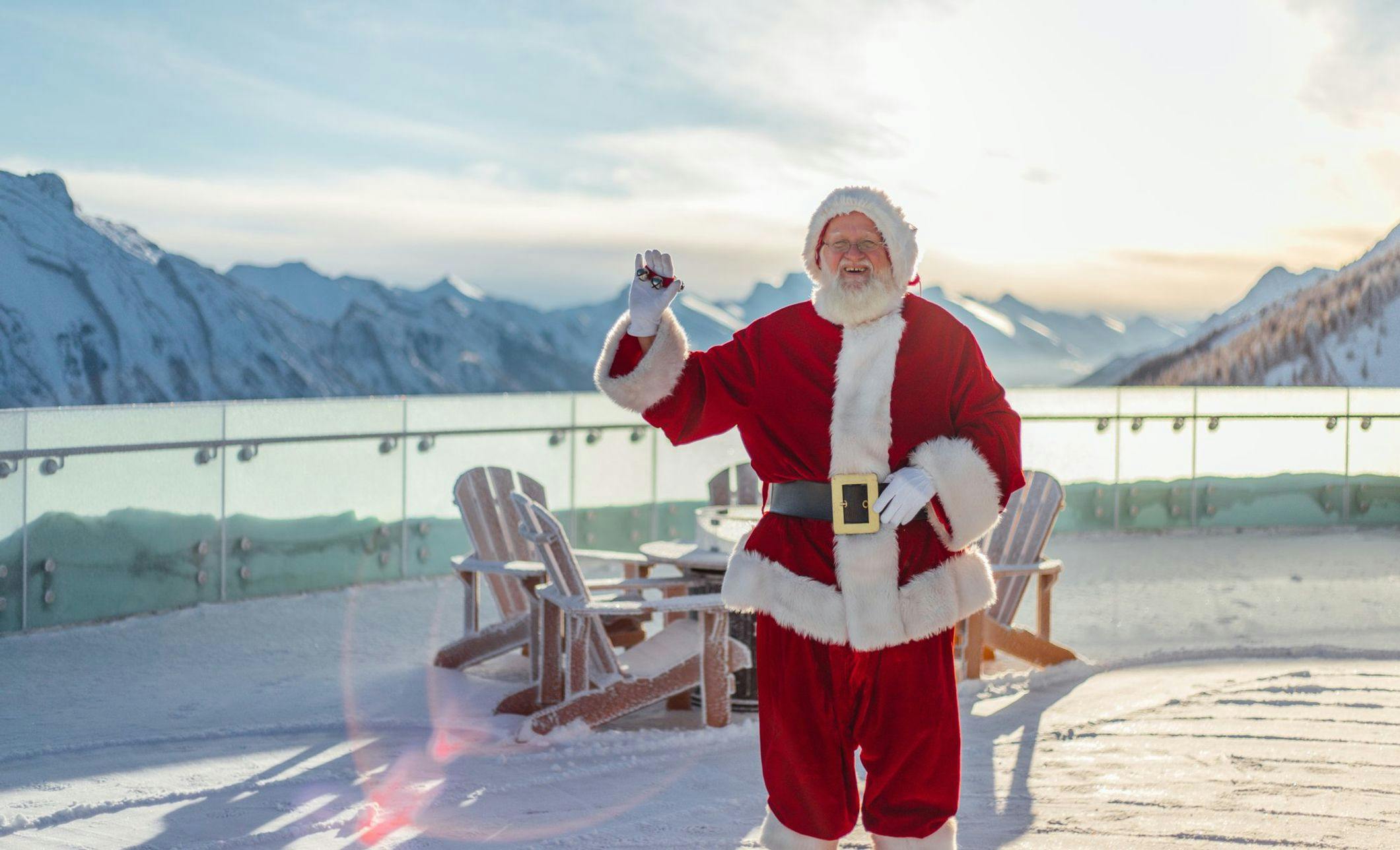 Santa Claus poses on a viewing platform on a bright winter day with mountaintops visible in the background