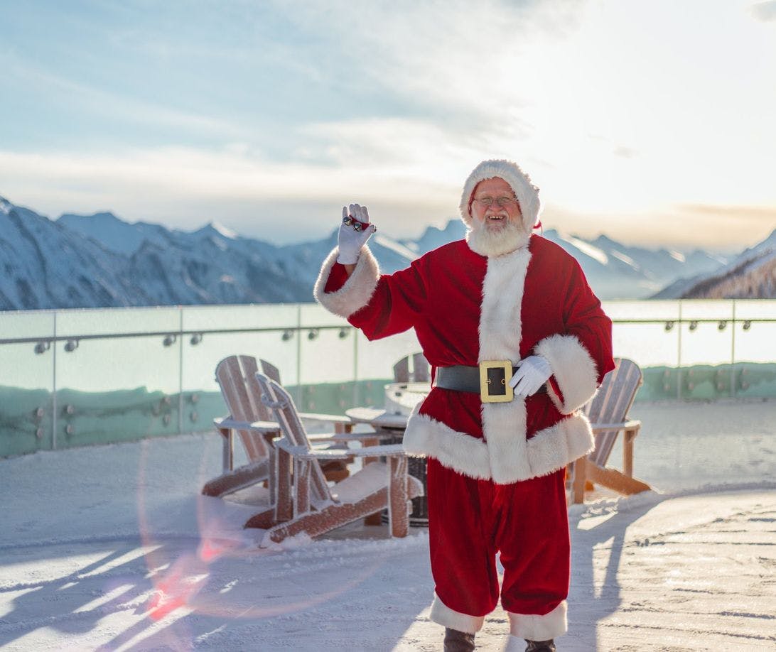 Santa Claus poses on a viewing platform on a bright winter day with mountaintops visible in the background