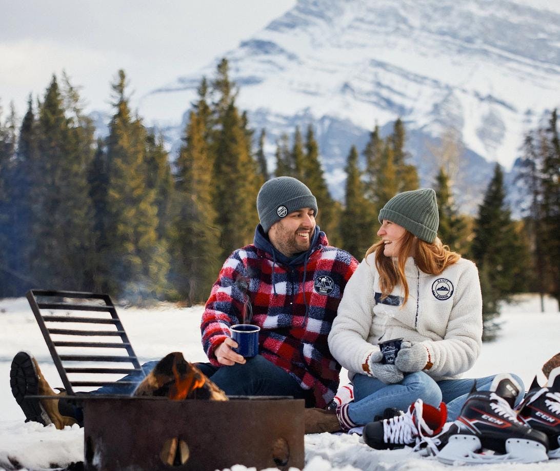 A couple sitting in front of a fire on a blanket while surrounded by snow. Holding cups of hot drinks and hockey skates beside them