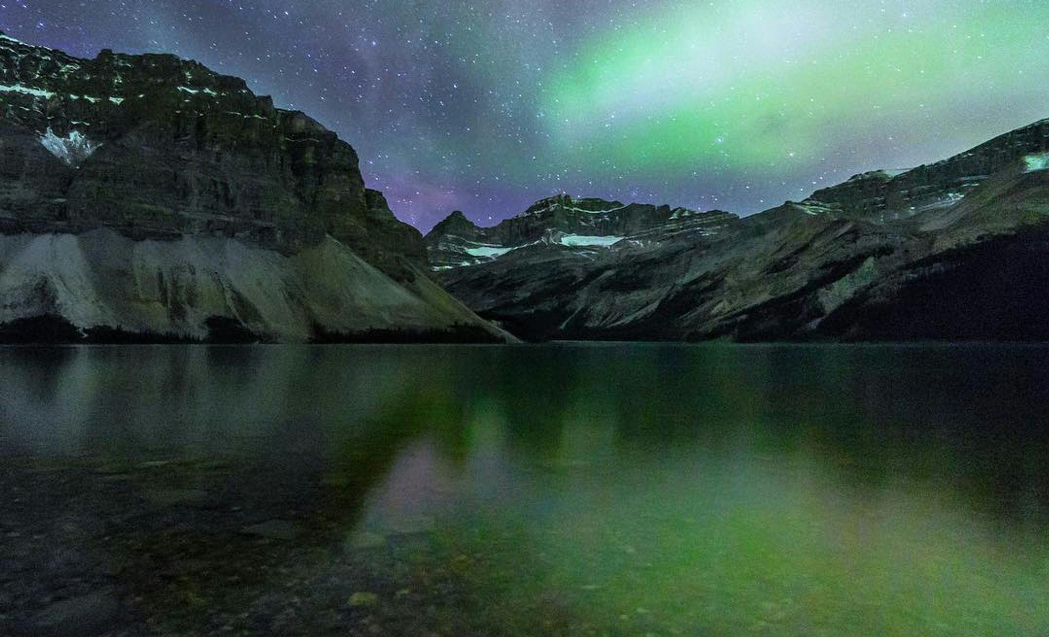 Colourful aurora and stars shine over mountaintops with Bow Lake in the foreground