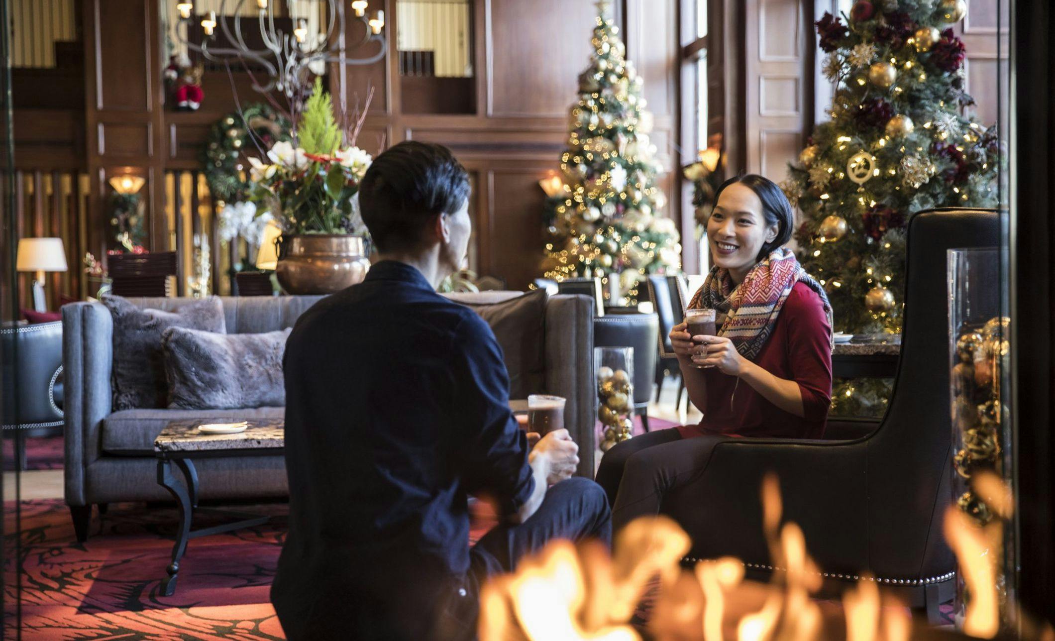A couple enjoys warm drinks next to an indoor fireplace in a lobby filled with lit Christmas trees
