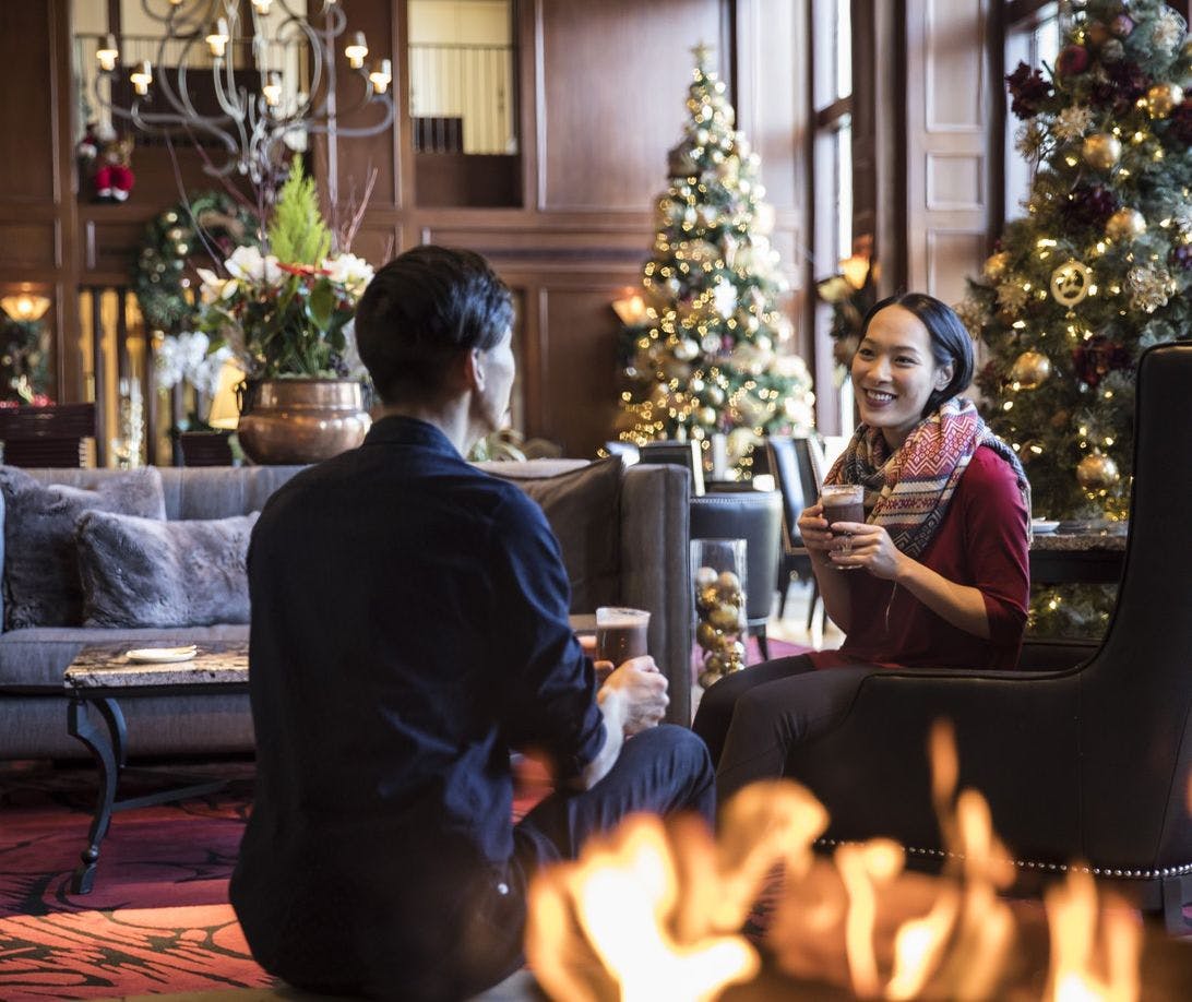A couple enjoys warm drinks next to an indoor fireplace in a lobby filled with lit Christmas trees