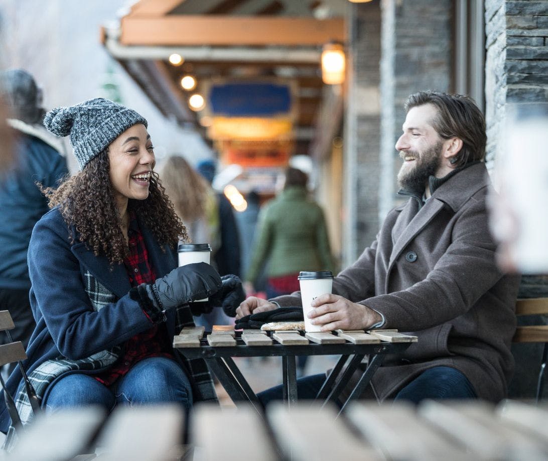 Couple sitting at an outdoor table wearing cozy mittens and toques and drinking coffee