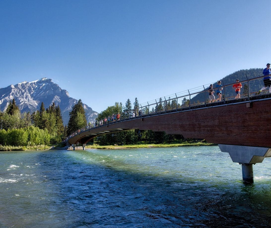 Runners cross a bridge over a turquoise river on a sunny summer day with mountains in the background