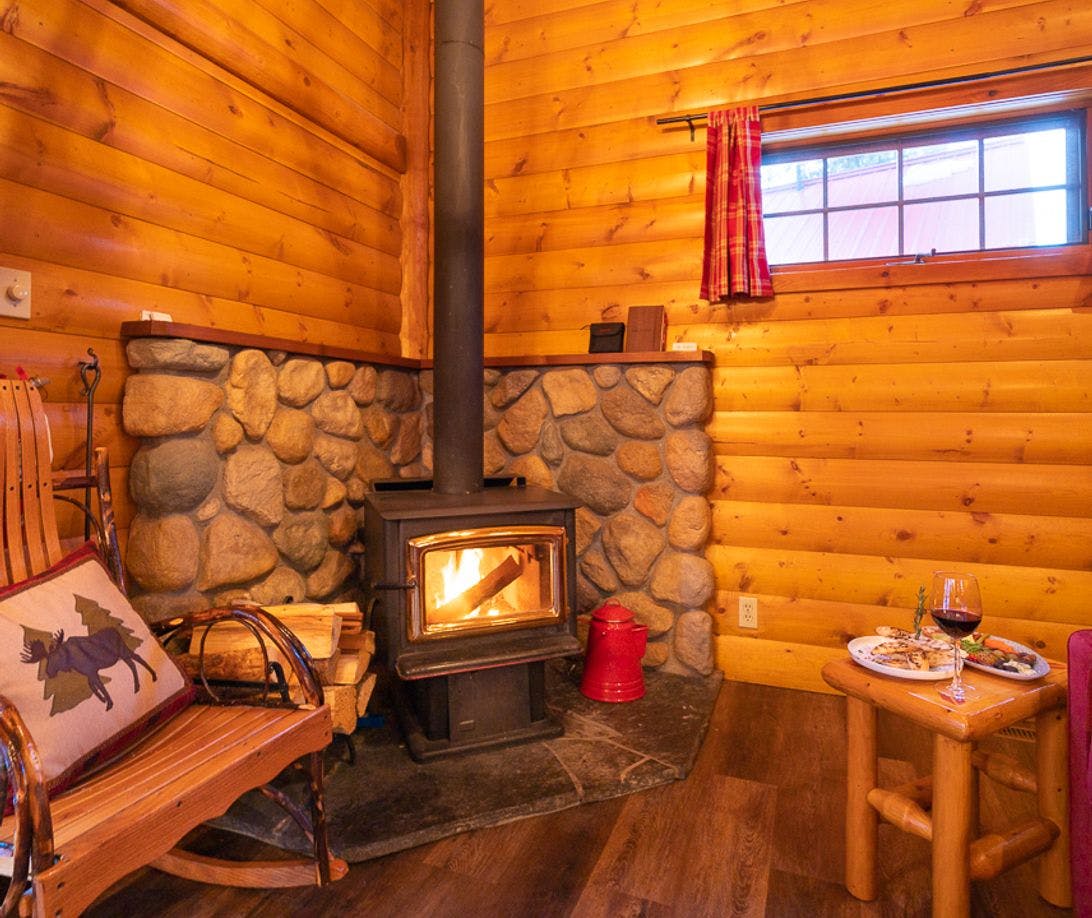 The interior of a cozy wooden cabin is lit with an indoor fireplace and there is wine and food on a table in front of the leather sofa