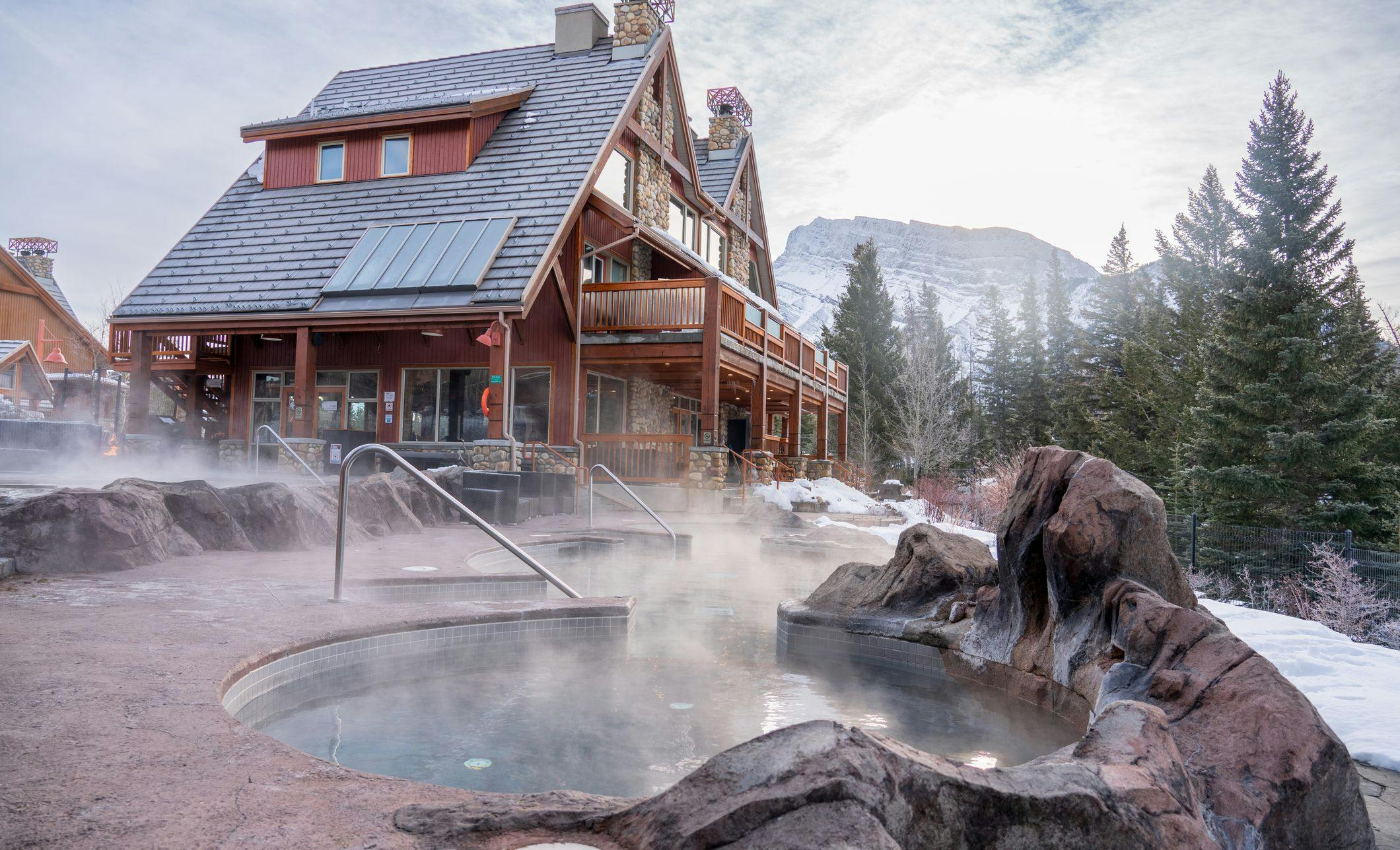 A lodge with an outdoor hot tub in front and snowy mountains surrounding