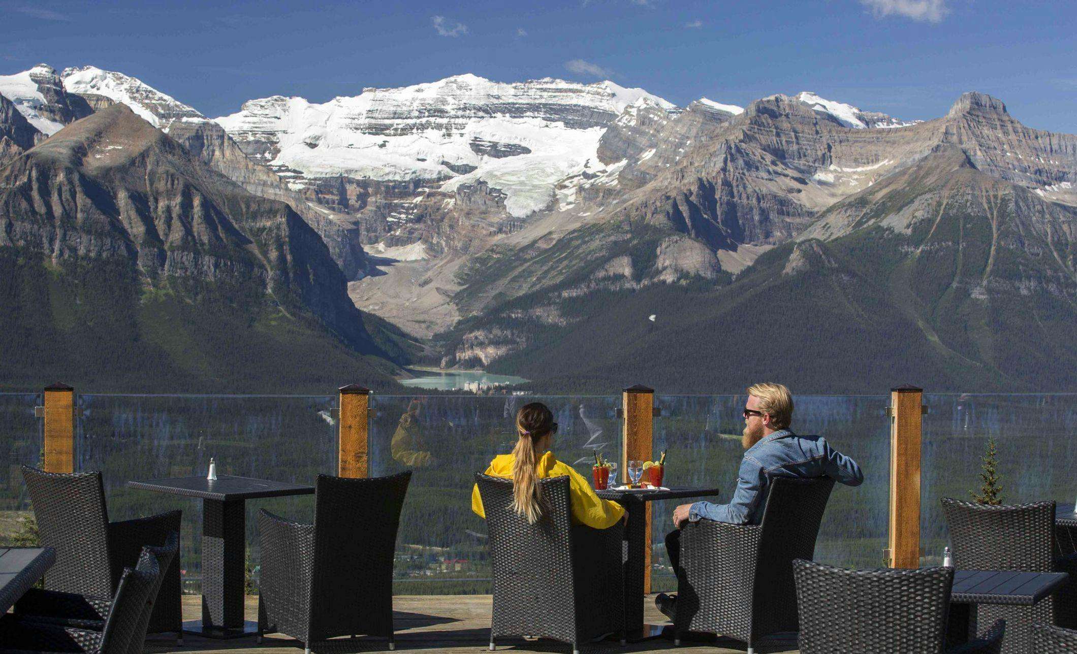 A couple sits at a table on a patio high up in the alpine overlooking Lake Louise and Victoria Glacier in the distance