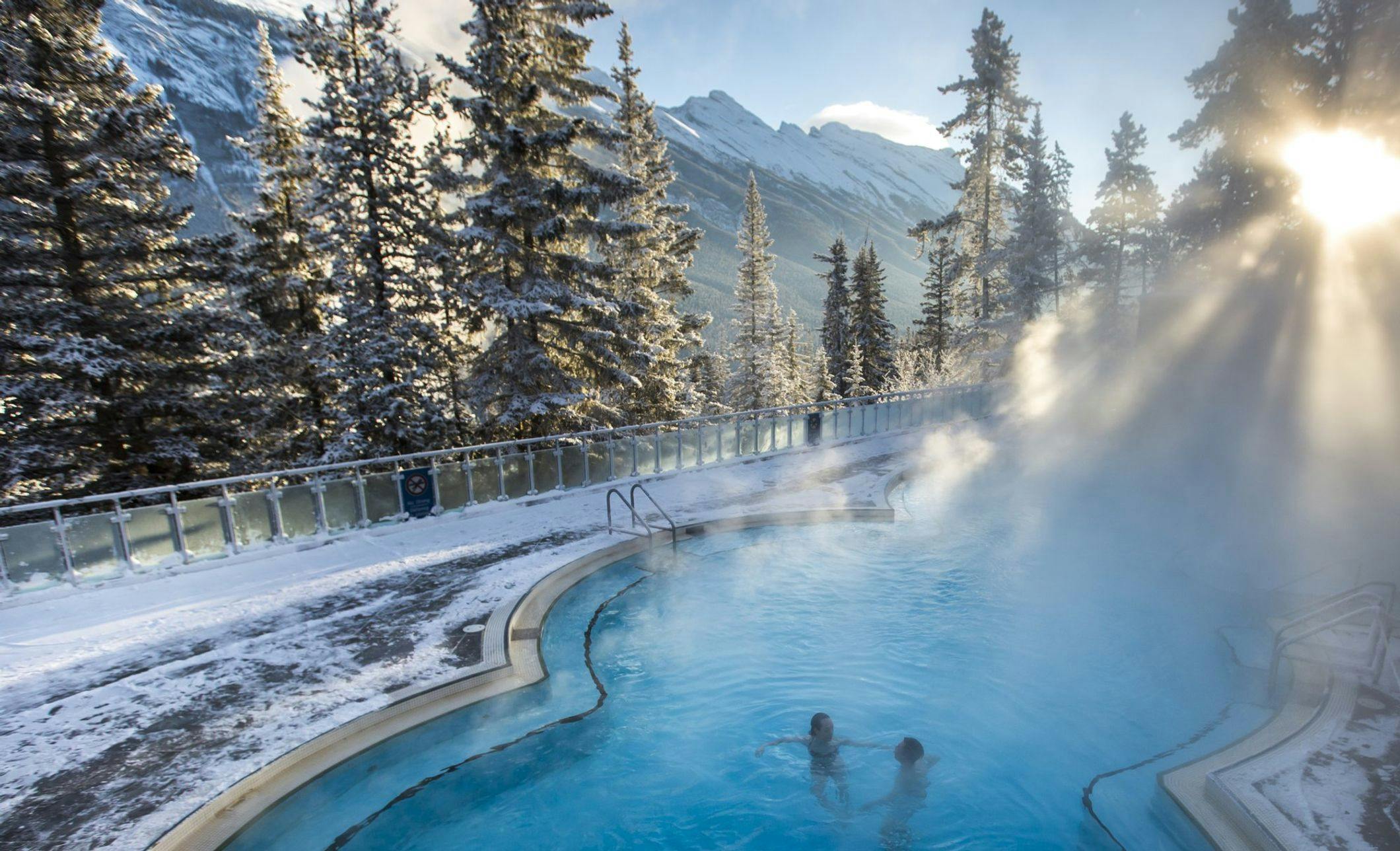 A couple soaking in an outdoor hot spring pool surrounded by snowy mountains and trees