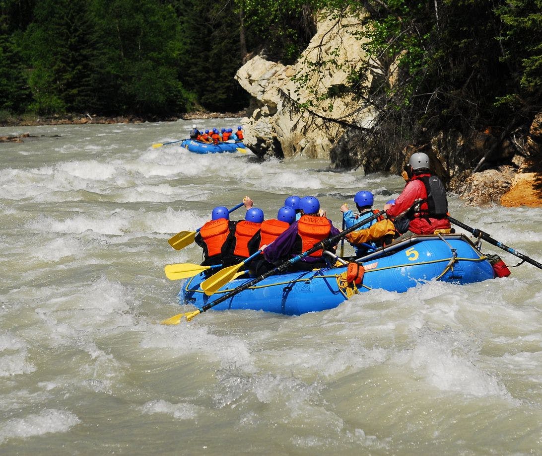 A group of rafters on a large blue raft navigate through rapids while smiling and being splashed with water