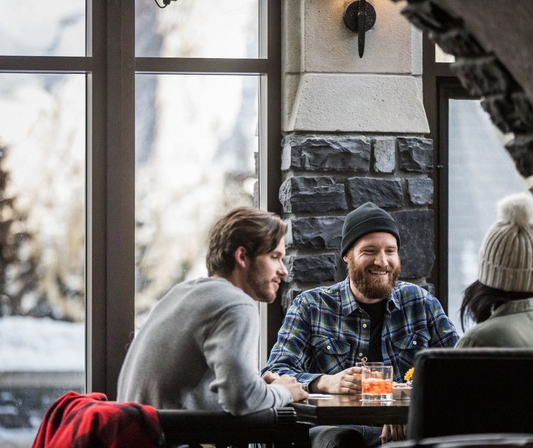 A group of three friends enjoy cocktails at a wooden table as fresh snow outside can be seen through the window. They are all wearing toques and layers after being out in the cold