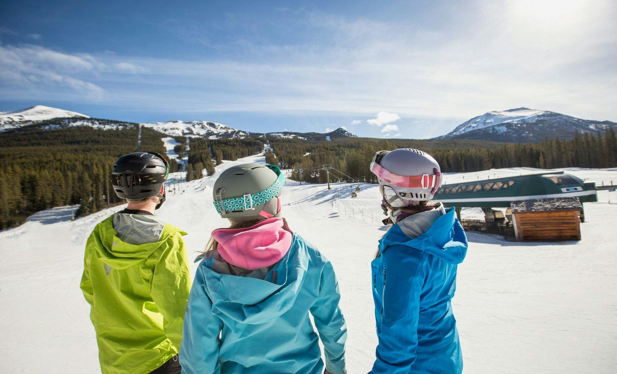 A small group of skiers/snowboarders stand together looking down at the ski terrain on a beautiful day with clear blue skies