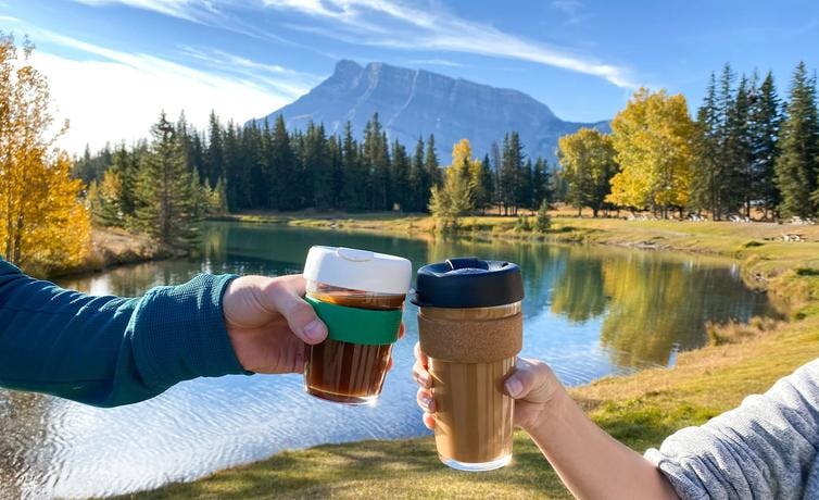 Two coffee cups "cheers-ing" at Cascade Ponds. Rundle Mountain and Fall colours in the background