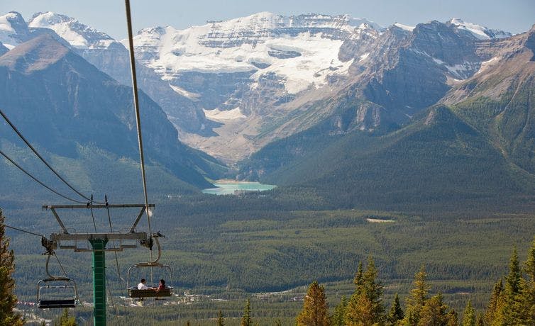 A couple travels down a mountain in an open chairlift while taking in the view of Lake Louise and Victoria Glacier in the distance