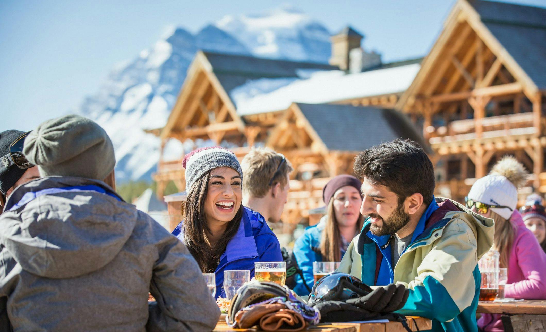 A group of friends enjoy a meal and drinks at an outdoor table at a ski resort. You can see the ski chalet and mountains in the background
