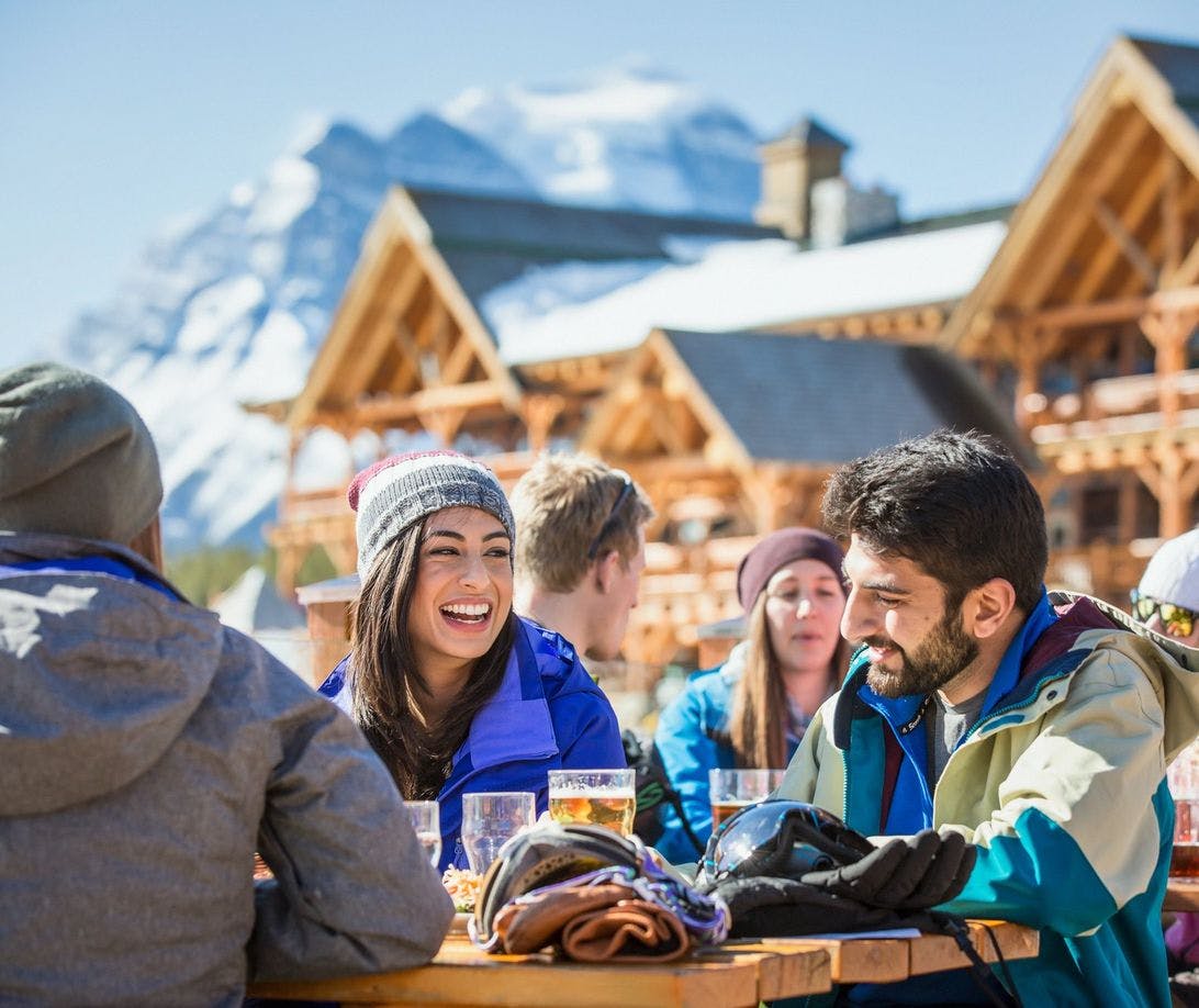 A group of friends enjoy a meal and drinks at an outdoor table at a ski resort. You can see the ski chalet and mountains in the background