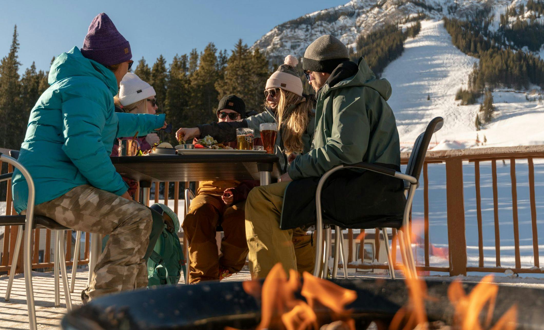 Friends sit around an outdoor table and a nearby outdoor fire pit with ski runs and snow in the background on a sunny day