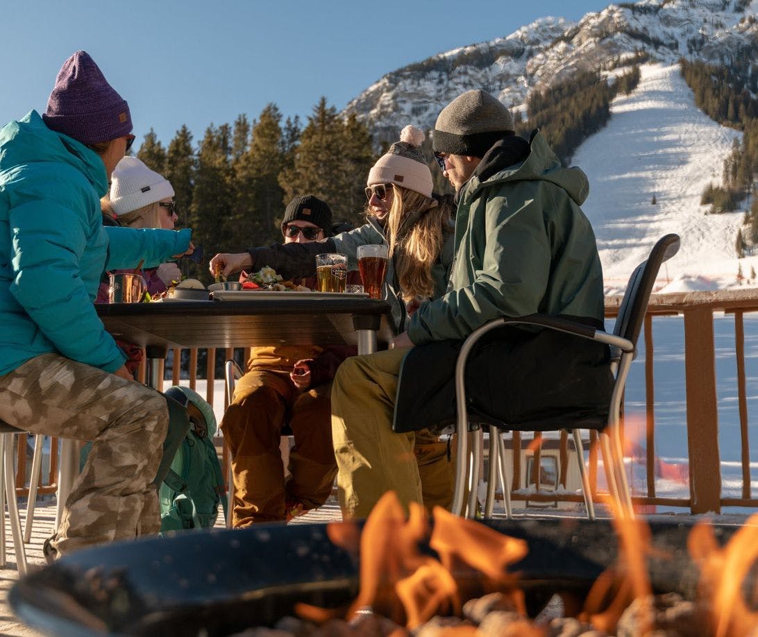 Friends sit around an outdoor table and a nearby outdoor fire pit with ski runs and snow in the background on a sunny day