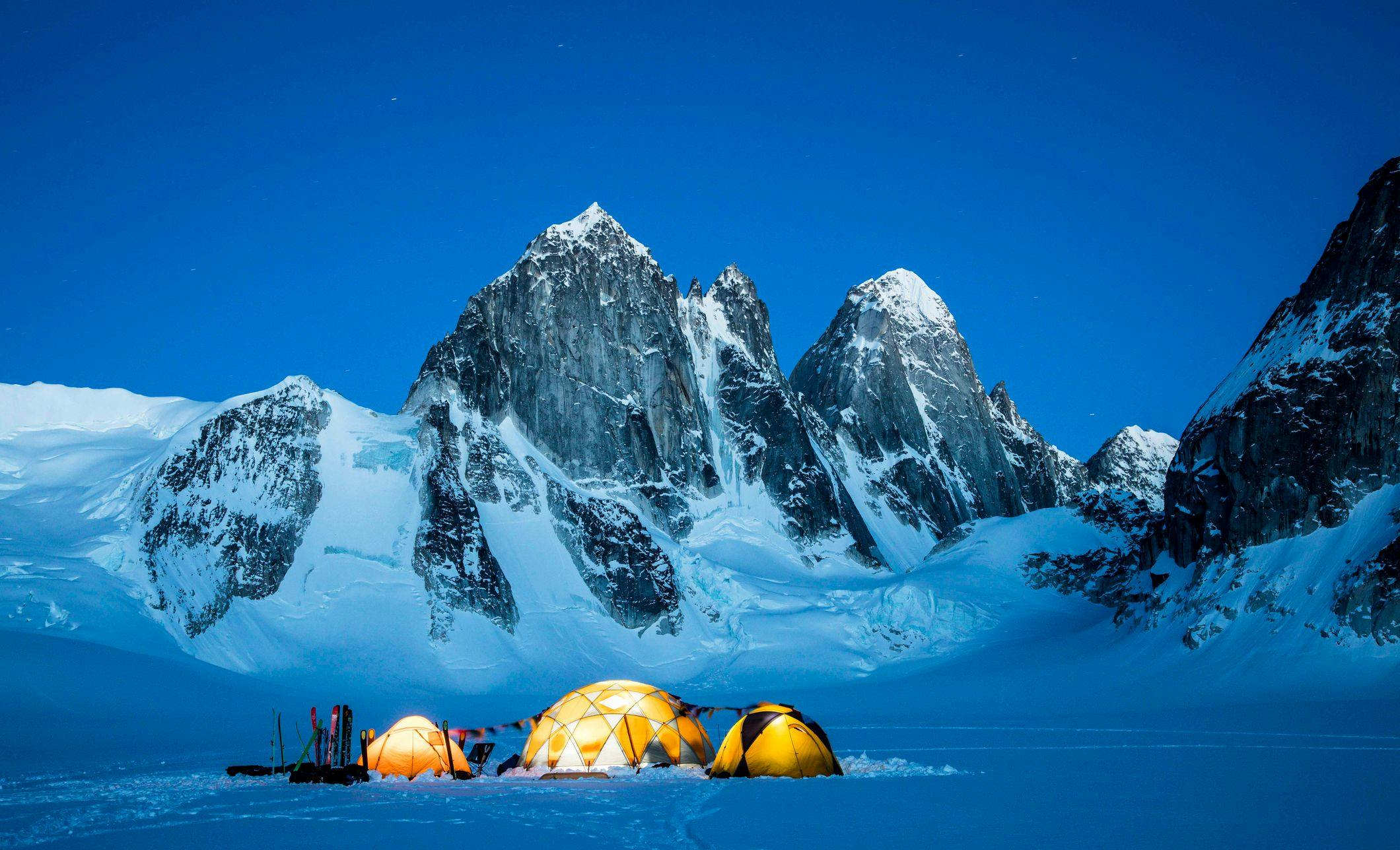Three lit up domes on a vast snowy field with skis outside and mountains in the background